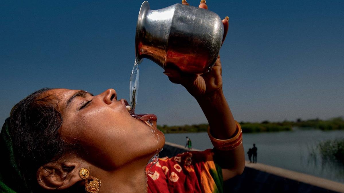 A woman drinking water from a steel pot beside a reservoir in rural India